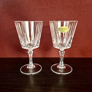Pode incluir: Duas taças de vinho de cristal transparente com design facetado. Cada taça tem uma haste e uma base redonda. Uma taça tem um rótulo que diz "Cristal d'Arques". As taças estão sobre uma superfície castanha escura, fundo bordô.