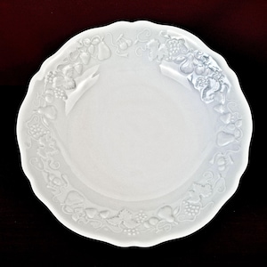 May include: A white ceramic plate with a decorative, scalloped edge. The rim is adorned with a raised relief pattern of fruits and vines. The plate is set against a dark background, emphasizing its bright white colour and detailed design.
