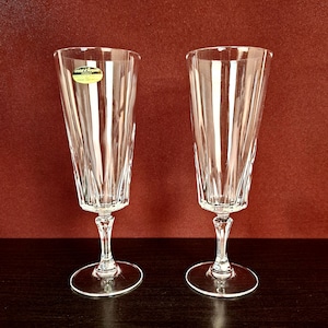 Pode incluir: Duas taças de champanhe transparentes com linhas verticais gravadas. Cada taça tem uma base redonda e uma haste longa e fina. Uma pequena etiqueta dourada é visível em uma taça. As taças estão sobre um fundo vermelho escuro.