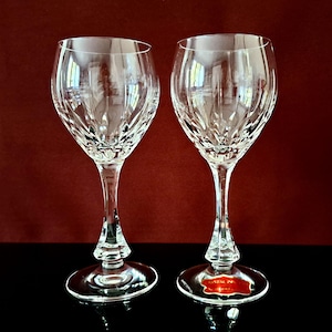 Könnte beinhalten: Zwei klare Weingläser aus Kristall mit aufwendigen Schliffmustern. Die Gläser haben lange Stiele und runde Füße. Auf dem Fuß eines Glases ist ein rot-weißes Etikett zu sehen. Der Hintergrund ist dunkelrot.