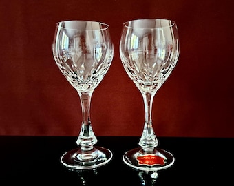May include: Two clear crystal wine glasses with intricate cut patterns. The glasses have long stems and round bases. A red and white label is visible on the base of one glass. The background is a dark red.