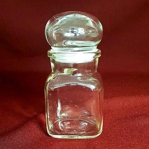 Peut inclure: Un pot en verre transparent avec un couvercle rond en verre. Le pot a une forme carrée et est vide.