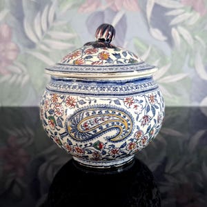 May include: A round, lidded ceramic jar with a detailed floral and paisley design. The jar features a white base with blue, red, and yellow floral patterns. The lid has a dark brown knob. The jar is likely for decorative or storage purposes.