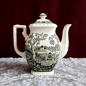 Può includere: Una teiera in ceramica bianca con un design floreale verde e bianco. La teiera ha un coperchio e un beccuccio. È appoggiata su un tovagliolo di pizzo bianco.