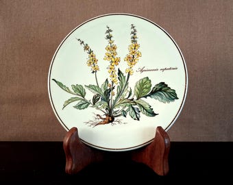 May include: A decorative plate featuring a botanical illustration of a yellow-flowered plant with green leaves and the text "Agrimonia eupatoria." The plate has a white background with a brown rim and is displayed on a wooden stand.