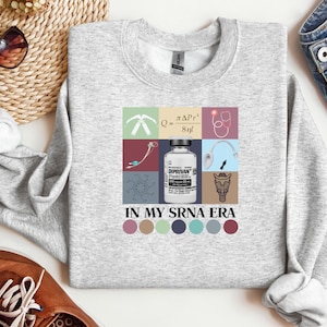 May include: A grey sweatshirt with a graphic design featuring a medical syringe, a stethoscope, a chemical formula, a bottle of medication labelled "Diprivan", and a cartoon animal. The text "In My SRNA Era" is printed below the graphic.