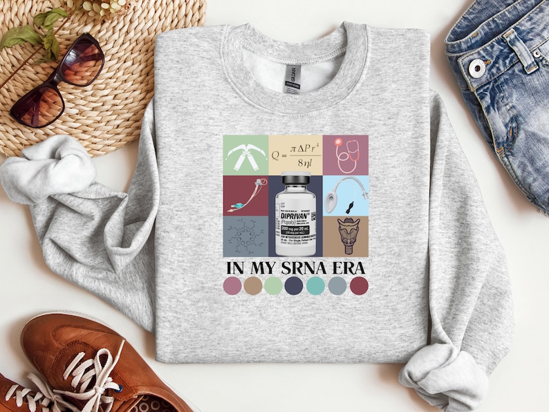 May include: A gray sweatshirt with a graphic design featuring a medical syringe, a stethoscope, a chemical formula, a bottle of medication labeled "Diprivan", and a cartoon animal. The text "In My SRNA Era" is printed below the graphic.