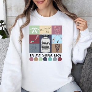 May include: White sweatshirt with a colorful graphic design featuring medical symbols, a vial of medicine, and the text "IN MY SRNA ERA". The graphic is surrounded by a row of six colorful circles.