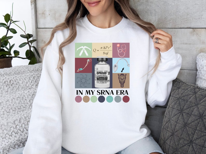 May include: White sweatshirt with a colorful graphic design featuring medical symbols, a vial of medicine, and the text "IN MY SRNA ERA". The graphic is surrounded by a row of six colorful circles.