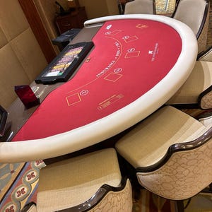 May include: A blackjack table with a red felt surface and white padded edges. The table has gold text and card symbols. Several upholstered chairs surround the table. The table is in a casino setting.