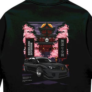May include: A black hooded sweatshirt featuring a graphic on the back. The design displays a black sports car parked in front of a traditional red temple surrounded by pink cherry blossoms. The text reads RECKLESS DIVISION and 無謀な分割.