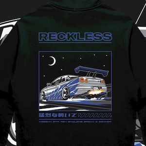 May include: Black hoodie with a graphic of a blue and silver sports car with a spoiler, flames, and the word "RECKLESS" above the graphic. The background is a night sky with a moon and stars.