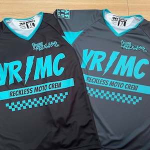 May include: Two black and turquoise youth motocross jerseys with the text "VR/MC Reckless Moto Crew" and "Young Reckless" printed on the front.