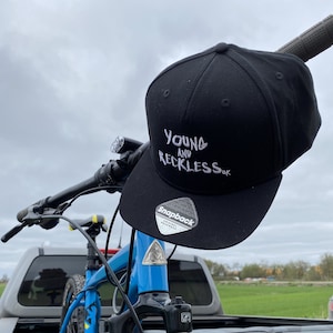 Puede incluir: Una gorra de béisbol negra y blanca con el texto "Young and Reckless UK" bordado en la parte delantera. La gorra está sentada en la parte trasera de una camioneta con una bicicleta de montaña azul.