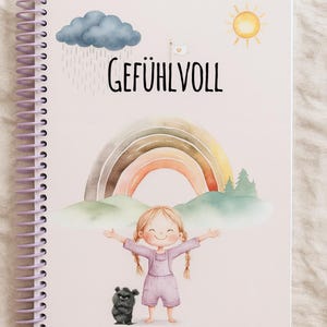 Könnte beinhalten: Spiralgebundenes Notizbuch mit einer Illustration. Auf dem Cover sind ein lächelndes Kind, ein Regenbogen, eine Sonne, eine Regenwolke und das Wort "GEFÜHLVOLL" abgebildet. Ein kleines, schwarzes, pelziges Wesen steht neben dem Kind.