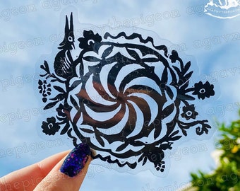 Könnte beinhalten: Ein schwarz-weißer Aufkleber mit einem floralen Design und einem wirbelnden Muster in der Mitte. Der Aufkleber befindet sich auf einem transparenten Hintergrund und wird von einer Hand gehalten.