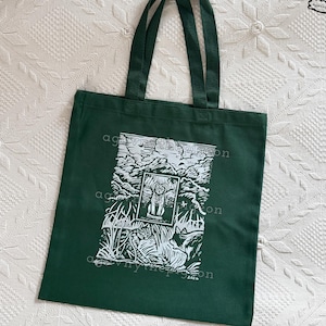 Peut inclure: Un sac fourre-tout vert fonc&eacute; avec un motif s&eacute;rigraphi&eacute; blanc repr&eacute;sentant un chat assis dans une fen&ecirc;tre encadr&eacute;e, entour&eacute; d'un paysage d'arbres et d'herbe. Le texte "agovnythepigeon" est imprim&eacute; au centre et en haut &agrave; droite.