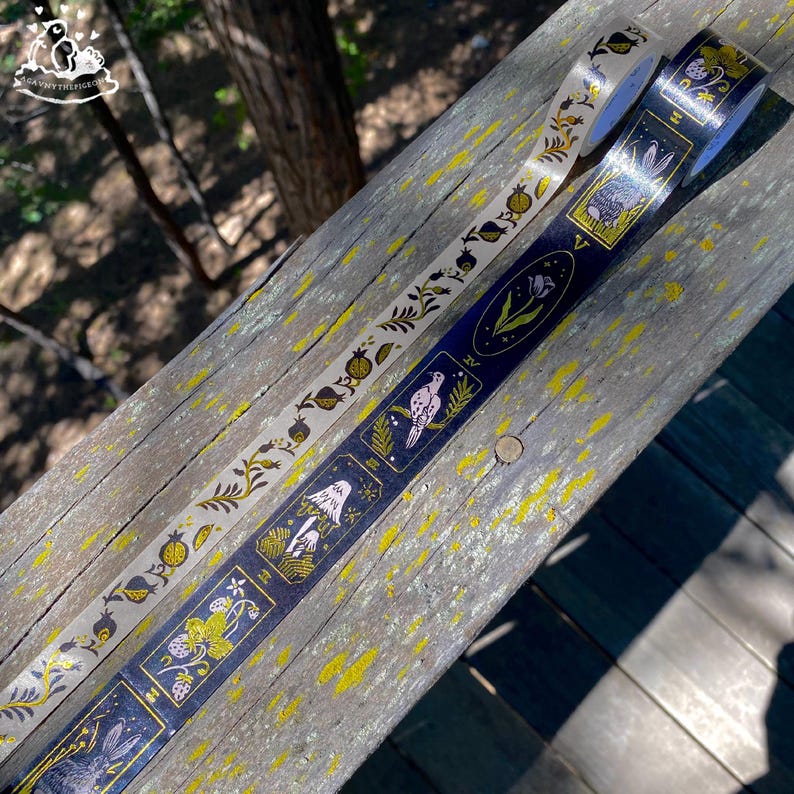 K&ouml;nnte beinhalten: Zwei dekorative Washi-Tapes mit Naturmotiven. Ein Band ist cremefarben mit einem sich wiederholenden Muster aus Granat&auml;pfeln und Bl&auml;ttern. Das andere ist marineblau mit Illustrationen von V&ouml;geln, Pilzen und Kaninchen. Beide B&auml;nder haben goldene Akzente.