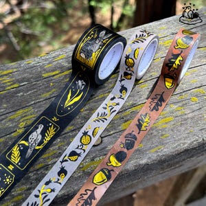K&ouml;nnte beinhalten: Drei dekorative Washi-Tape-Rollen mit Naturmotiven. Eine Rolle ist schwarz mit einem Hasen- und Blumenmuster, eine andere grau mit Granat&auml;pfeln und die dritte braun mit Eicheln und Bl&auml;ttern. Alle haben goldene Akzente.