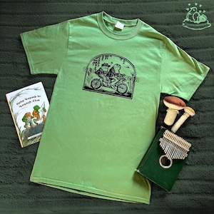 May include: A light green t-shirt featuring a black illustration of two frogs riding a bicycle. A book with text in a foreign language and a small kalimba are next to the shirt. Two decorative mushroom figures are also visible.