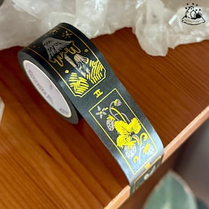 K&ouml;nnte beinhalten: Schwarzes Washi-Tape mit goldenen und gelben Illustrationen von Pilzen und Erdbeeren. Das Klebeband tr&auml;gt den Text "THE WASHI STATION" an der Seite und liegt auf einer Holzoberfl&auml;che.