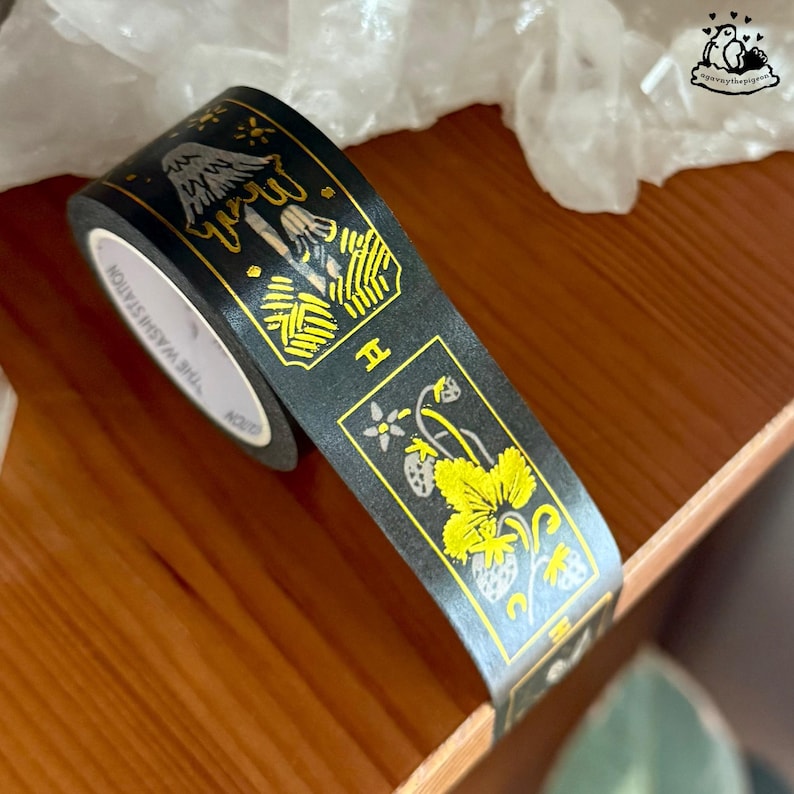 K&ouml;nnte beinhalten: Schwarzes Washi-Tape mit goldenen und gelben Illustrationen von Pilzen und Erdbeeren. Das Klebeband tr&auml;gt den Text "THE WASHI STATION" an der Seite und liegt auf einer Holzoberfl&auml;che.