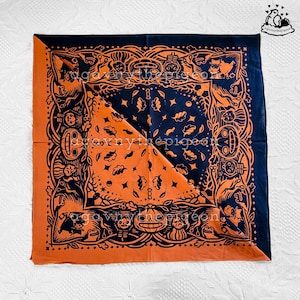 Puede incluir: Un pañuelo cuadrado con un diseño diagonal dividido en negro y naranja. La tela muestra un estampado detallado de calabazas de Halloween, esqueletos y hojas de otoño. El texto "agavnythepigeon" está superpuesto en el centro de la tela.