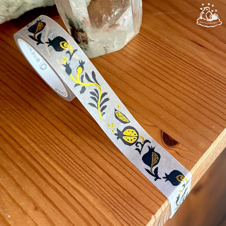 K&ouml;nnte beinhalten: Dekoratives Washi-Tape mit einem sich wiederholenden Muster aus schwarzen und gelben Granat&auml;pfeln und floralen Elementen auf transparentem Hintergrund. Das Band ist gerollt und teilweise abgerollt. Das Band liegt auf einer Holzoberfl&auml;che.