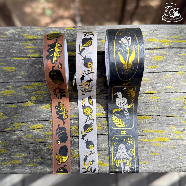 K&ouml;nnte beinhalten: Drei dekorative Washi-Tape-Rollen mit Naturmotiven. Eine Rolle zeigt Eicheln und Eichenbl&auml;tter auf braunem Hintergrund. Eine andere hat Granatapfel- und Blumenmuster auf wei&szlig;em Hintergrund. Die dritte Rolle zeigt einen Vogel und einen Pilz auf schwarzem Hintergrund.