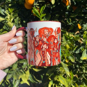 May include: A white ceramic mug with a red rim and handle features a colorful illustration of women in traditional clothing. The women are depicted in a circle, with their hands raised in a celebratory gesture. The mug is a great gift for anyone who appreciates art and culture.