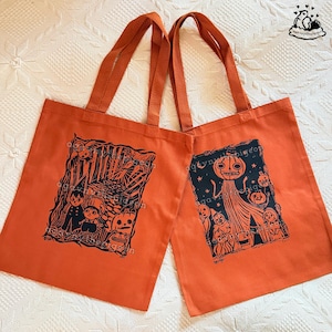 May include: Two orange tote bags with black screen-printed illustrations. The left bag features a forest scene with characters and a pumpkin. The right bag displays a tall pumpkin-headed figure surrounded by smaller figures and stars.