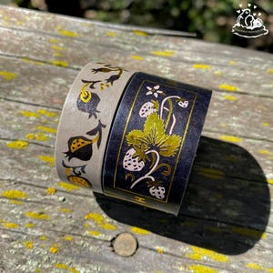 K&ouml;nnte beinhalten: Zwei Rollen dekoratives Washi-Tape. Eine Rolle ist grau mit einem schwarzen und goldenen Blumendesign. Die andere Rolle ist schwarz mit einem goldenen und wei&szlig;en Erdbeer- und Blattdesign. Das Klebeband liegt auf einer Holzoberfl&auml;che.