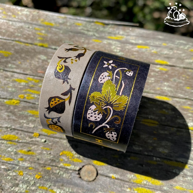 K&ouml;nnte beinhalten: Zwei Rollen dekoratives Washi-Tape. Eine Rolle ist grau mit einem schwarzen und goldenen Blumendesign. Die andere Rolle ist schwarz mit einem goldenen und wei&szlig;en Erdbeer- und Blattdesign. Das Klebeband liegt auf einer Holzoberfl&auml;che.