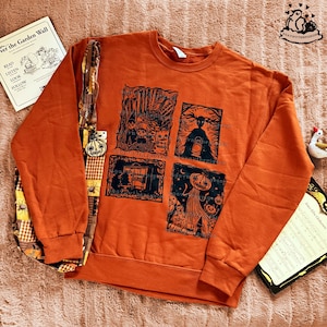 Könnte beinhalten: Ein orangefarbenes Sweatshirt mit Rundhalsausschnitt und vier schwarz-weißen Illustrationen zum Thema Halloween. Das Sweatshirt wird mit einer gemusterten Tragetasche und einem Buch mit dem Titel "Over the Garden Wall" präsentiert.