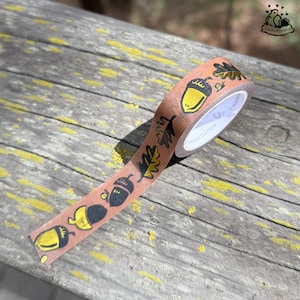K&ouml;nnte beinhalten: Eine Rolle Washi-Tape mit braunem Hintergrund und einem sich wiederholenden Muster aus gelben Eicheln und schwarzen Bl&auml;ttern. Das Tape liegt auf einer verwitterten Holzoberfl&auml;che.