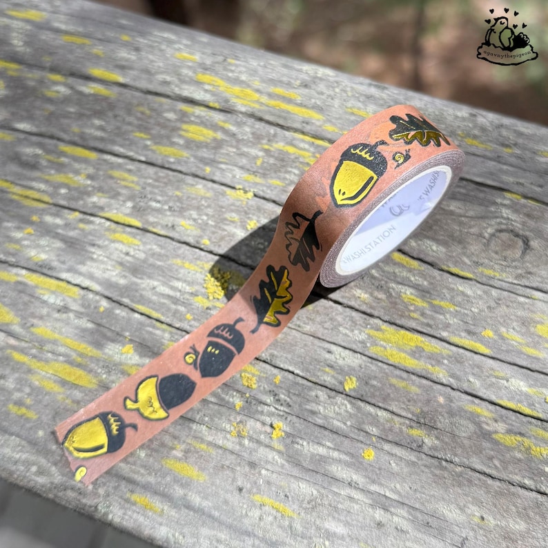 K&ouml;nnte beinhalten: Eine Rolle Washi-Tape mit braunem Hintergrund und einem sich wiederholenden Muster aus gelben Eicheln und schwarzen Bl&auml;ttern. Das Tape liegt auf einer verwitterten Holzoberfl&auml;che.