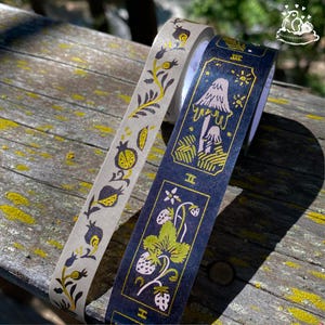 K&ouml;nnte beinhalten: Zwei dekorative Washi-Tape-Rollen. Eine Rolle ist hellgrau mit einem goldenen Blumendesign. Die andere ist marineblau mit einem goldenen Pilz- und Erdbeerdesign. Das Klebeband befindet sich auf einer verwitterten Holzoberfl&auml;che.