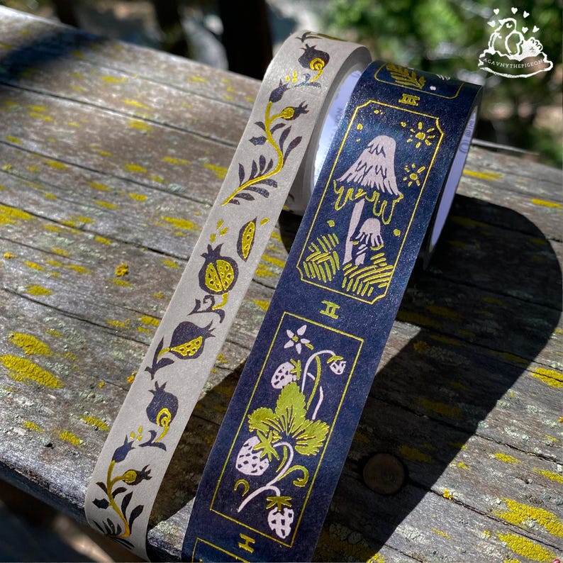 K&ouml;nnte beinhalten: Zwei dekorative Washi-Tape-Rollen. Eine Rolle ist hellgrau mit einem goldenen Blumendesign. Die andere ist marineblau mit einem goldenen Pilz- und Erdbeerdesign. Das Klebeband befindet sich auf einer verwitterten Holzoberfl&auml;che.