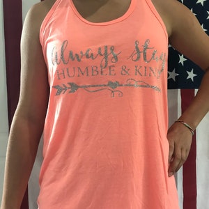 Puede incluir: Un top de tirantes de color rosa coral con el texto "Always Stay Humble & Kind" en letras plateadas con purpurina y un gráfico de flecha y corazón.