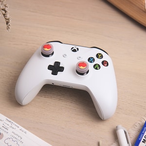 Peut inclure: Une manette Xbox blanche avec des capuchons de boutons en forme de pomme rouge et jaune. La manette est sur une surface en bois clair.