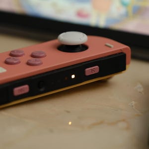 Op de afbeelding: Een koraalkleurige gamecontroller met een witte joystick en roze knoppen. De controller heeft een zwarte basis met de letters "SL" en een klein lampje. De controller is ontworpen voor gaming.