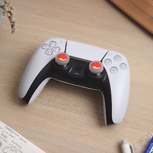 May include: A white and black PlayStation 5 DualSense controller with two red and yellow apple-shaped thumbstick caps.