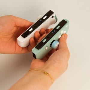 May include: Two game controllers, one white and one mint green, are held in hands. Each controller has black accents, buttons, and a thumb grip. The thumb grips are light blue with a snowflake design. The controllers are designed for gaming.