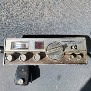 May include: A silver and brown Realistic TRC-424 CB radio with a digital display and several knobs and buttons. The radio has a black microphone attached to it.