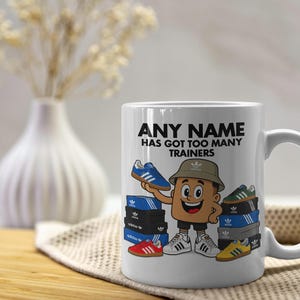 May include: A white ceramic mug featuring a cartoon character wearing a bucket hat, surrounded by stacks of shoeboxes and various trainers. The text printed on the mug reads, ANY NAME HAS GOT TOO MANY TRAINERS, with the Adidas logo on the boxes.