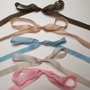 Peut inclure: Cinq rubans de soie de différentes couleurs, chacun noué en un nœud. Les couleurs comprennent le vert olive, le beige, le bleu clair, l'argent et le rose. Les bords des rubans sont effilochés, ajoutant une touche rustique. Parfait pour l'artisanat, l'emballage de cadeaux ou les accents décoratifs.