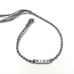 May include: Braided bracelet with the name "NICO" spelled out in white letter beads. The bracelet is made of red, white, and blue threads, with a braided design. The bracelet is on a white background.
