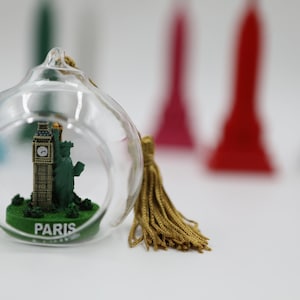 May include: A glass ornament with a gold tassel features a miniature replica of Big Ben and the Statue of Liberty. The base of the ornament reads "Paris" in black lettering.