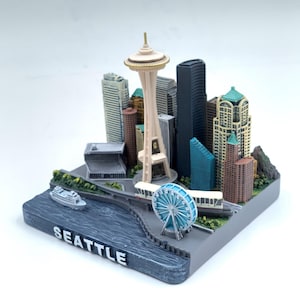 Könnte beinhalten: Miniatur-Souvenir der Skyline von Seattle mit Space Needle, Wolkenkratzern, einem Riesenrad und einer Fähre. Der Sockel ist grau mit dem weißen Wort "SEATTLE". Die Gebäude sind in verschiedenen Farben gehalten.