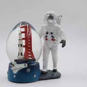 May include: A snow globe with a red and white rocket and a red tower inside. An astronaut figurine stands next to the snow globe. The base of the snow globe is blue with white stars and a white space shuttle.
